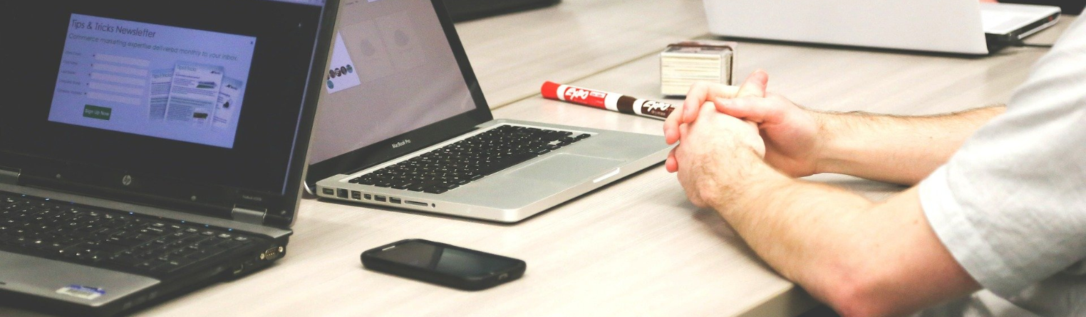 Two laptops and a smarpthone on a desk and a human sitting at the computer with classped hands.